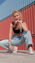 Blonde Caucasian woman wearing denim pants crouches in front of shipping containers