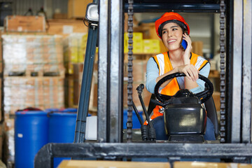 female worker talking by smartphone and driving on forklift in warehouse storage