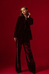 Fashionable confident woman wearing dark red velvet suit blazer, wide leg trousers, posing on red...
