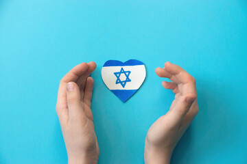 child's hands and a paper-cut drawn Israeli flag in the shape of a heart on a blue background....