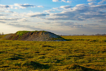 A grassy field with a large mound, under a sky with scattered clouds.