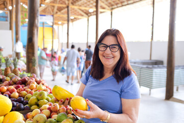 A joyful and smiling brazilian mature woman at a market, holding two papayas, illustrates the...