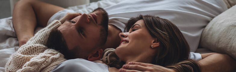 Banner relationships. Beautiful happy loving young couple relaxing in bed, looking at each other. Cozy home atmosphere, tenderness, closeness. Embracing, kissing. Lazy weekend, slow living concept - obrazy, fototapety, plakaty