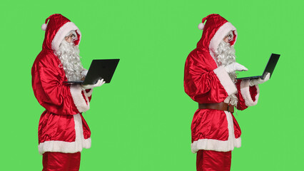 Santa claus man on online videocall waving at laptop screen to discuss with people about gifts, standing over greenscreen background. Person in red festive suit on teleconference call.