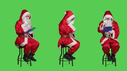 Saint nick writing notes on clipboard while he sits on chair dressed in red costume and white beard, full body greenscreen. Father christmas character holding papers in studio.