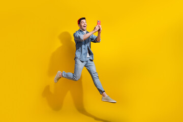 Fototapeta na wymiar Full length photo of impressed funky man dressed jeans shirt jumping high chatting modern device empty space isolated yellow color background