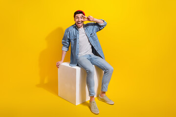 Full length photo of excited positive man dressed jeans shirt sitting white cube showing v-sign cover eye isolated yellow color background
