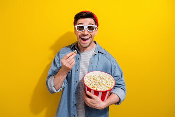 Photo of excited cheerful man dressed jeans shirt enjoying 3d film eating pop corn isolated yellow...