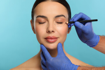 Doctor with pencil preparing patient for cosmetic surgery operation on light blue background,...