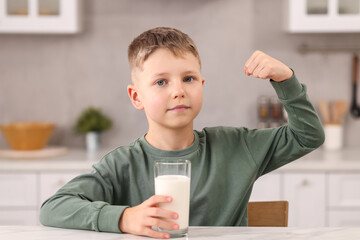 Cute boy with glass of fresh milk showing his strength at white table in kitchen