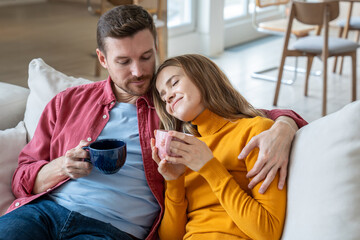 Pleased couple enjoying lazy weekend resting at home drinking coffee together on cozy sofa. Husband...