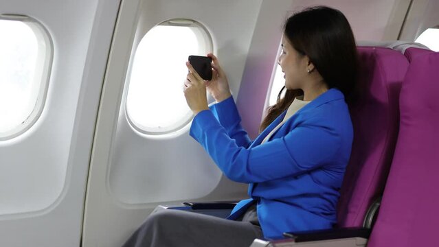 Attractive Asian businesswoman sitting in airplane cabin playing with mobile phone and taking pictures from window.