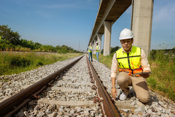 male Asian engineer wearing a hard hat sits and inspects the quality of the railroad tracks, and...