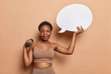 Horizontal shot of dark skinned fitness woman raises dumbbell trains muscles shows white speech...