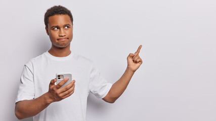 Serious dark skinned man holds smartphone and points index finger at blank space presses lips wears casual tshirt shows place for your advertisement isolated over white background. Hmm look at this