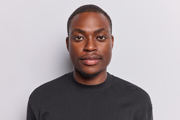 Portrait of handsome dark skinned man looks directly at camera has serious expression full lips concentrated gaze wears casual black t shirt isolated over white background poses for making photo - obrazy, fototapety, plakaty