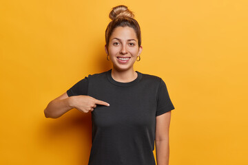Waist up shot of positive young woman with hair bun points at blank space of black t shirt for your...