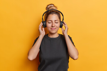 Horizontal shot of pleased calm young woman wears headphones on ears enjoys listening music keeps eyes closed dressed in casual black t shirt isolated over yellow background. Favorite hobby concept