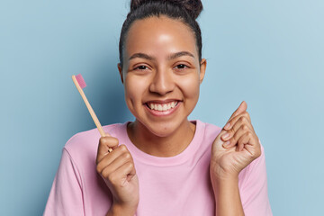 Smiling cheerful Latin woman with dark hair gathered in bun holds wooden toothbrush going to clean teeth smiles broadly clenches fist dressed in casual t shirt isolated over blue background.