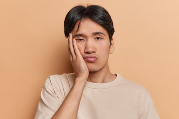 Portrait of displeased Japanese man has dull day keeps hand on cheek showing lack of interest feels...