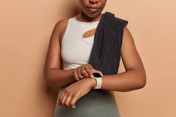 Cropped shot of unrecognizable active sportswoman checks fitness results on smartwatch dressed in activewear poses with towel on shoulder leads active lifestyle isolated over brown background