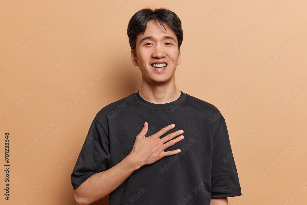 Wall mural photo of handsome dark haired young chinese man keeps hand on chest dressed in casual black t shirt  - Wall murals