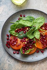 Olive-colored plate with sliced persimmon, fresh baby spinach and pomegranate seeds, vertical shot on a beige granite surface, middle closeup