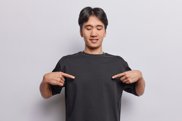 Mockup for design. Pleased dark haired man points at blank black t shirt shows place for your advertisement focused down isolated over white background. People clothing and promotion concept