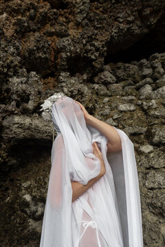 A girl covered with a veil poses in a crown of shells against the backdrop of a reef and rocks