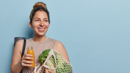 People and sporty lifestyle concept. Happy positive sporty woman with hair bun holds bottle of juice and net bag of fresh vegetables carries rolled karemat for fitness training isolated over blue wall