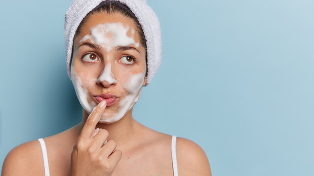 Studio shot of pensive European woman washes face with soap bubble keeps finger on lips concentrated aside wears wrapped bath towel on head isolated over blue background copy space for your promo
