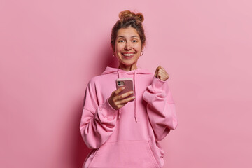 Cheerful overjoyed woman with hair bun clenches fist and rejoices success holds mobile phone in...