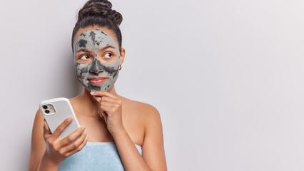 Thoughtful brunette Latin woman keeps hand on chin applies facial clay mask for skin treatment wrapped in towel holds mobile phone chats online isolated over white background copy space for promo