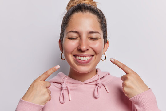 Horizontal shot of positive young European woman keeps eyes closed points both index fingers at toothy smile shows perfect white teeth dressed in casual hoodie isolated. Look at my brilliant smile