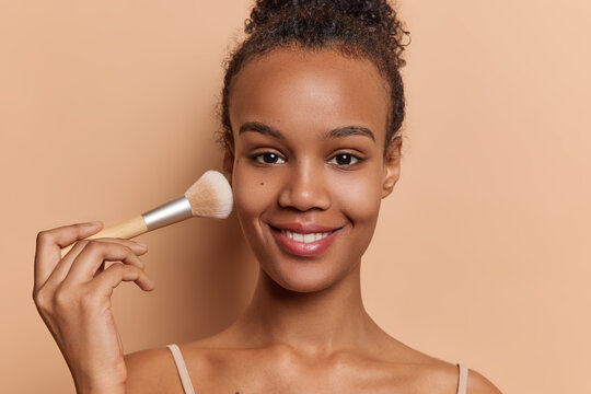 Smiling beautiful young woman with dark skin applies powder or foundation using cosmetics brush putting daily makeup after morning shower poses against brown background. Beauty routine concept.