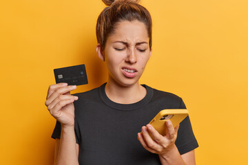Studio shot of young confused European lady holding and looking at smartphone paying online but seeing wrong card number standing in centre isolated on yellow background wearing black casual t shirt