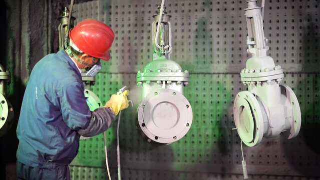A factory worker in a paint shop in a red helmet paints a pipeline tap with a spray gun