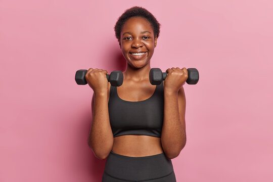 Active slim woman dressed in black cropped top and leggings lifts dumbbells being in good physical shape smiles pleasantly isolated over pink background doing biceps curl. Healthy lifestyle concept © Wayhome Studio