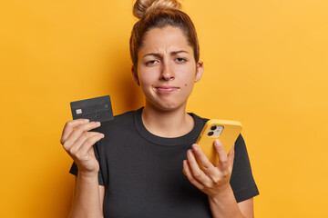 Photo of frustrated young woman holds mobile phone and credit card makes payment online feels...