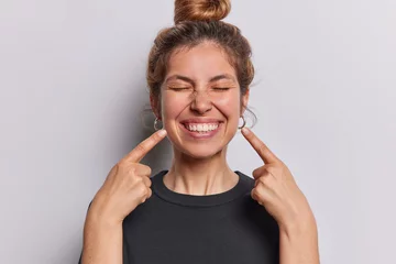 Fotobehang Photo of pleasant looking cheerful woman points index fingers at toothy smile shows wwell cared whie even teeth dressed in casual black t shirt isolated over whtie studio background feels happy © Wayhome Studio