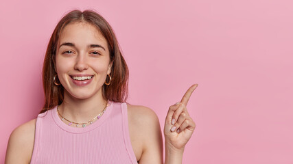 This way. Smiling brown haired young woman with happy expression points index finger on copy space for your promotional content wears casual t shirt isolated over pink background shows banner
