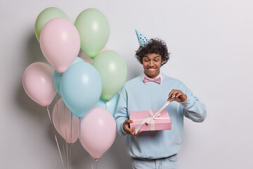 Positive curly Hindu man unwrapping birthday present with curious expession wears party hat blue pullover and jeans stands near bunch of inflated colorful balloons isolated over white background