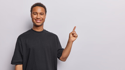 Handsome cheerful man smiles pleasantly dressed in casual black t shirt points index finger on...