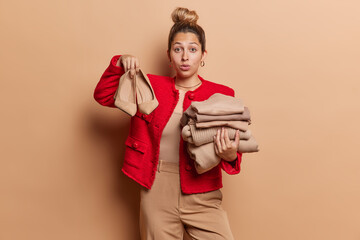 Indoor shot of impressed young woman poses with stack of clothing and high heeled shoes choosing footwear for her outfit wears stylish red jacket and trousers isolated over brown wall. Fashion concept