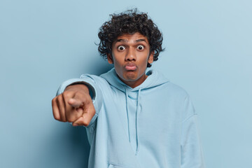 Shocked handsome curly haired Hindu man points index finger at camera notices something amazing...