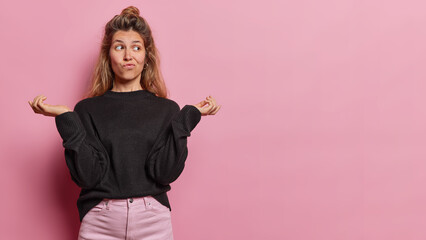 Uncertain clueless European woman spreads palms and shrugs shoulders purses lips concentrated aside dressed in casual clothes isolated over pink background copy space for your advertisement. - obrazy, fototapety, plakaty