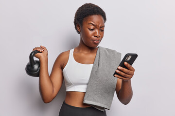 Sportswoman with dark skin lifts weight and focused at smartphone checks burned calories via...