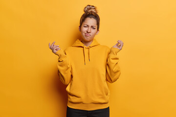 People emotions concept. Indoor photo of young confused European girl wearing yellow hoodie and black trousers standing in centre isolated spreading palms in hesitation not knowing what to do - obrazy, fototapety, plakaty