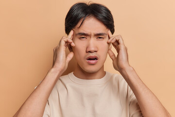 Photo of displeased Chinese man keeps hands on temples suffers from headache massages temples tries to concentrate or remember something dressed in casual t shirt isolated over brown background.