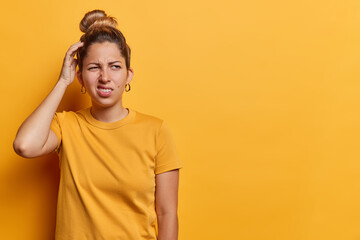 Waist up shot of European woman with hair bun scratches head and looks with bewilderment...